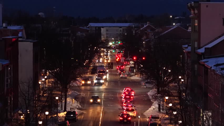 Busy main street at dusk with steady traffic and glowing lights, snow along sidewalks and rooftops. Urban winter scene with warm reflections and evening atmosphere in United States. Aerial rise zoom.