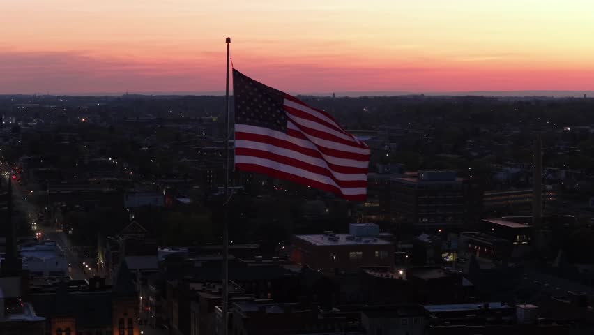 Waving American flag over Patriotic during sunset dusk. Aerial orbit shot. Pride and peaceful city in USA. Historic buildings and downtown in background. Stripes and stars of flag, detail shot.