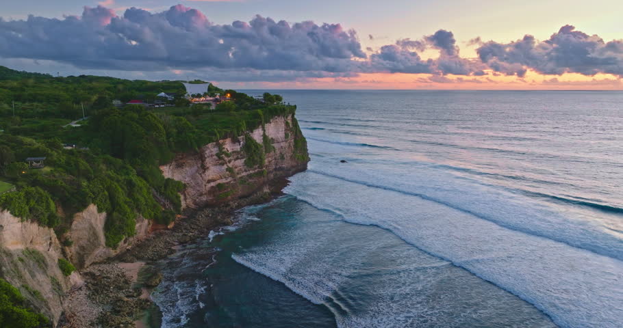 Tropical coastline creates a dramatic cliffside landscape, with green vegetation cascading down to the ocean, and waves breaking under a vibrant sunset sky. Bali travel. Aerial drone flight panorama
