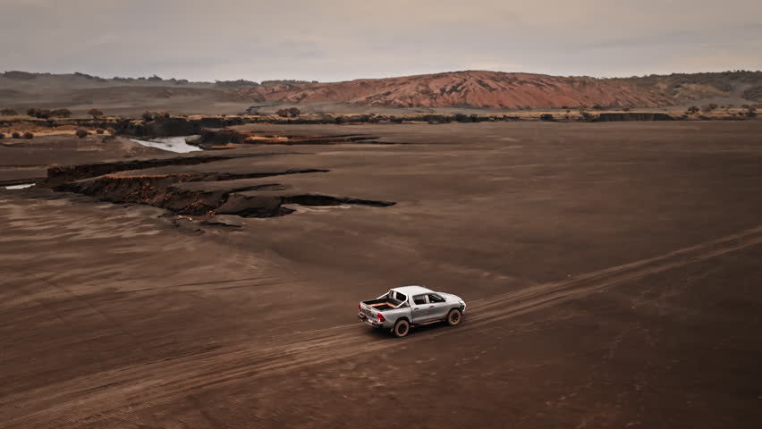 White pickup truck car driving volcanic landscape near Port Resolution on Tanna Island, dark ash, sand with eroded earth formations and a river flowing under overcast sky. Aerial view drone flight