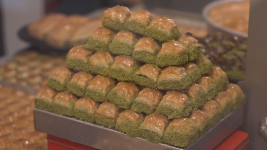 A mouth-watering 4K cinematic close-up of various traditional Turkish baklava types in a shop window. Features a red gift box labeled "ISTANBUL" among pistachio and walnut desserts.