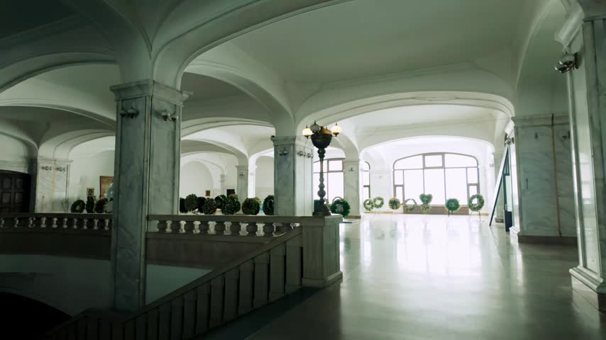 Bright hall within Holy Trinity Cathedral in Tbilisi, Georgia. Features marble columns, archways, large windows, and numerous floral wreaths arranged on display stands.