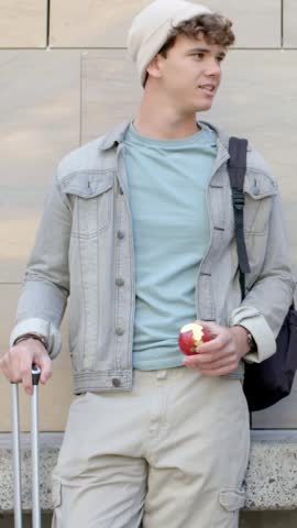 Vertical video: Man leaning in beanie with suitcase, biting apple and looking right on cue at plaza. Traveler, urban, outdoor, luggage, backpack, denim, vertical video