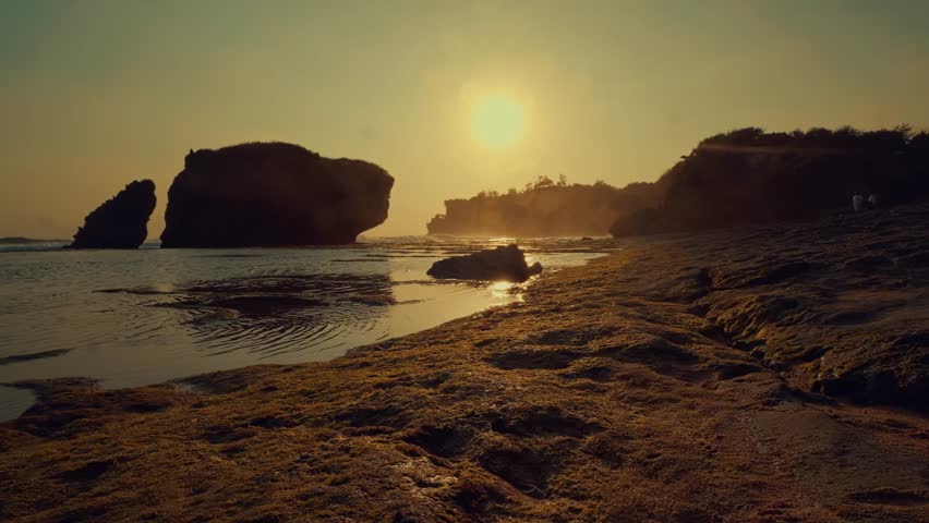 A serene coastal landscape featuring massive rock formations silhouetted against a radiant sunset. Low tide reveals textures on the wet sandy shore in a tranquil, cinematic scene.