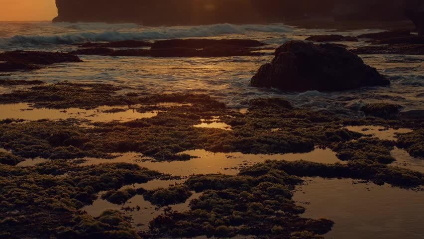A cinematic low angle shot of coastal rock formations covered in moss with gentle tide waves washing over a sunset landscape during the golden hour light.