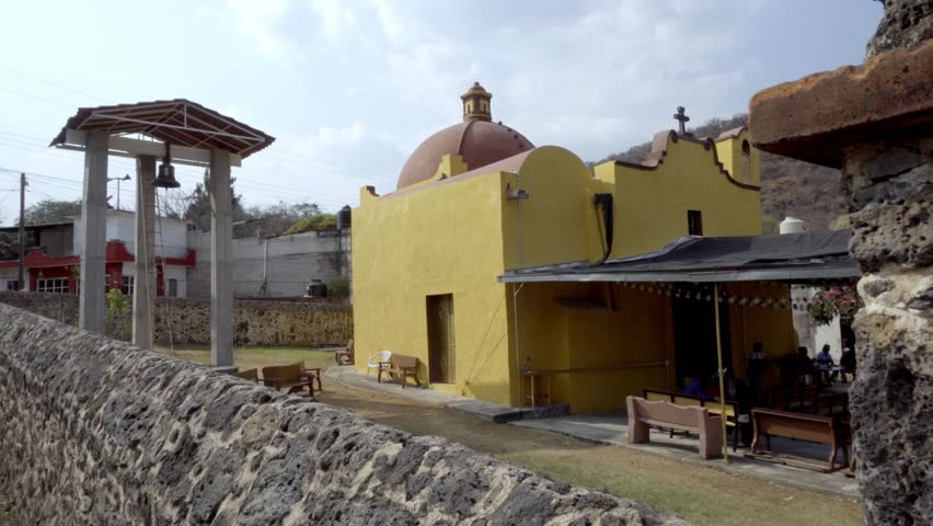 Small yellow church with dome and simple facade, bell structure nearby and stone wall in foreground, rural setting with benches and open space in daylight