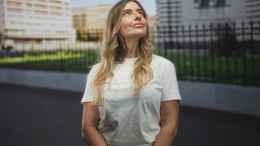 Woman in white t shirt gazes upward with closed eyes and slight smile, forearms exposed, standing on street by iron fence and city buildings; serene reflection.