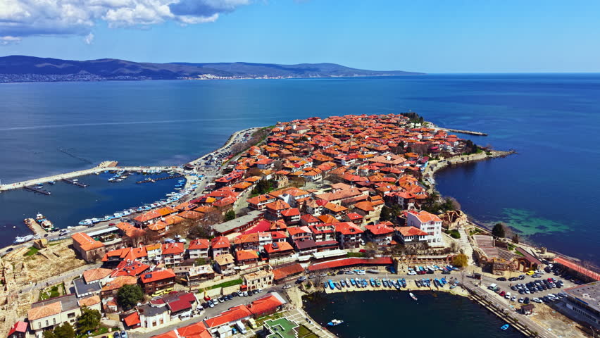 Visitors enjoy the views of Nesebar Old Town, featuring red roofs and historic buildings next to the blue sea. Boats are in the harbor as people stroll the streets by the water.