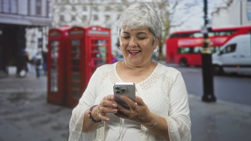 Woman holding smartphone with both hands on street by red telephone booth, looking up and smiling with bracelets and grey hair; surprise discovery.