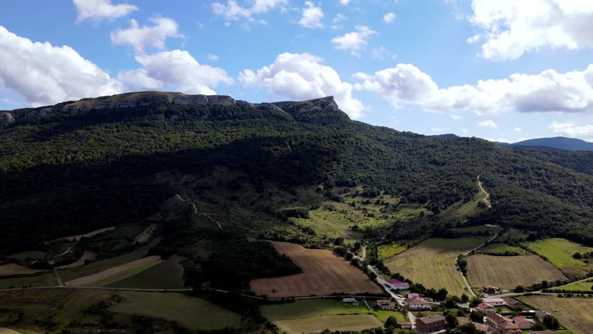 Cinematic drone shot approaching the picturesque village of San Roman (Alava) in Northern Spain. View of traditional houses, green fields, and a massive mountain ridge under a blue cloudy sky.