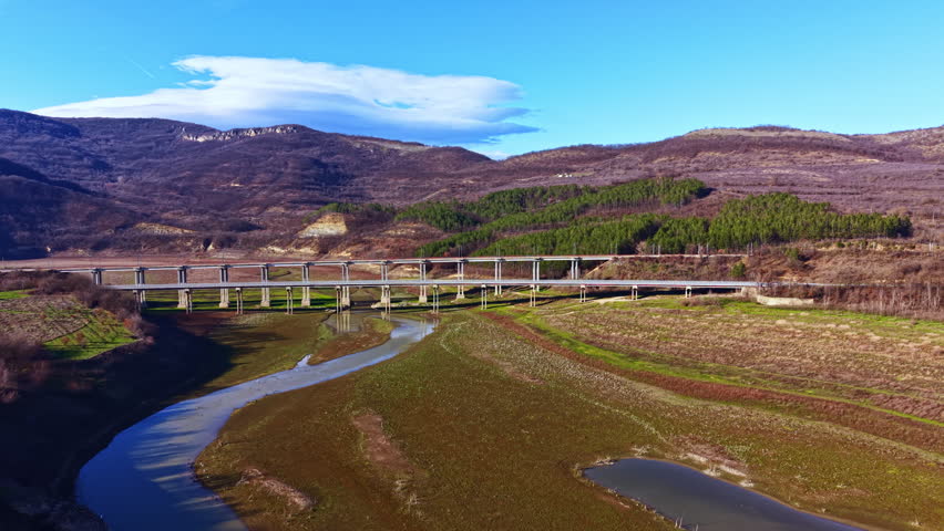 A bridge spans across a river in a valley. Hills rise in the background with patches of green trees. The landscape shows dry ground and few plants, while the sky is blue with some clouds.
