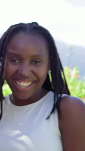 Vertical video: Laughing woman with locs responding to lens moving closer in sun, wearing white top. Outdoors, daylight, sunlit, vibrant, candid, portrait, vertical video