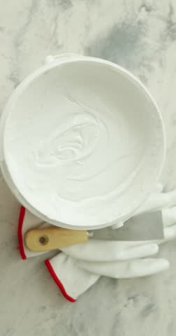 Bucket of plaster, putty knife and gloves on light textured table, flat lay. Camera spinning