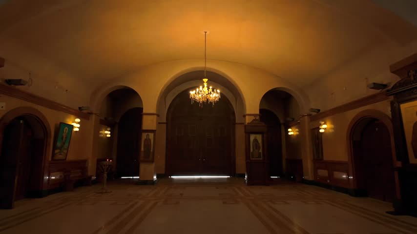 Expansive interior hall of Holy Trinity Cathedral, showcasing a grand chandelier illuminating the space. Features multiple arched doorways, a symmetrical layout, and large imposing wooden doors at ...