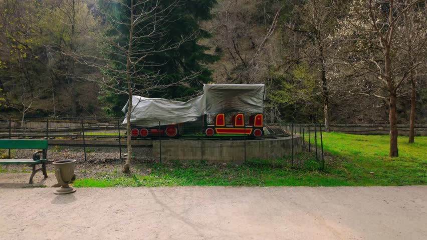 View of a small, covered amusement park train on its track, nestled among tall trees and vibrant green grass, with a park bench nearby in Borjomi, Georgia.