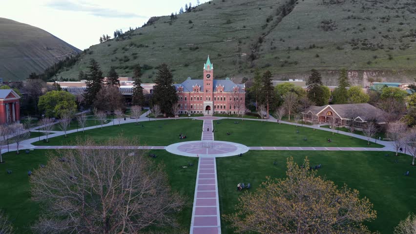 Historic university campus in Missoula, Montana, United States, with central lawn, academic buildings and surrounding hills under soft evening light. Calm educational atmosphere. Aerial drone approach