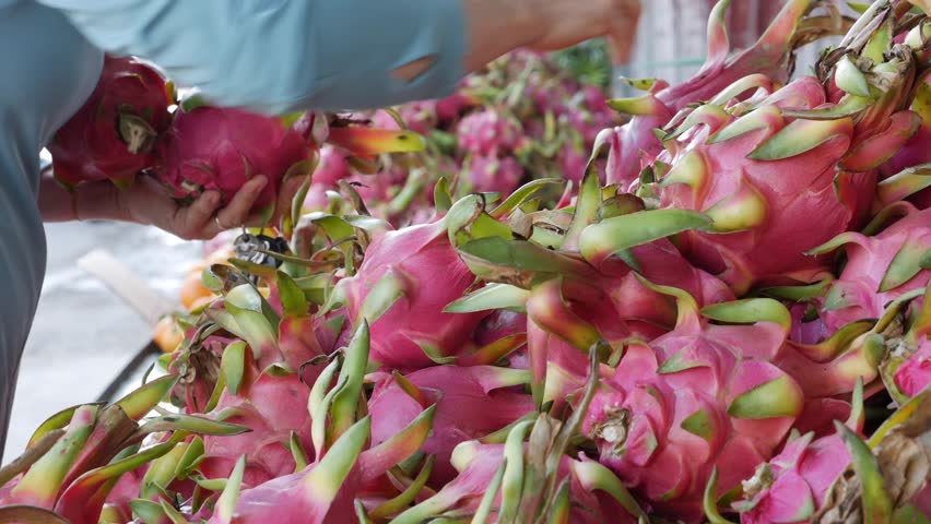 Hand selects dragon fruits from a pile on a market counter while fruits are arranged together on a stall display in an outdoor market setting. Market stall.