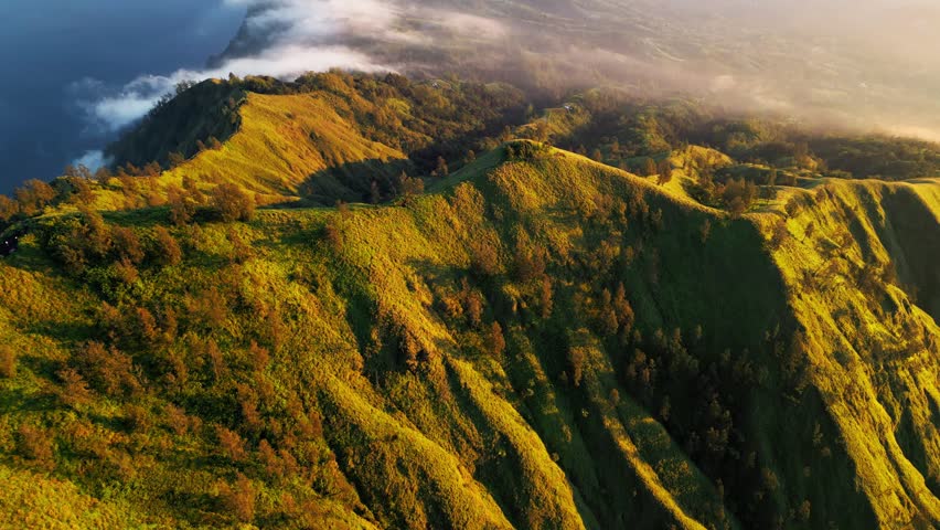 Mountain peak with steep grassy slopes and crater lake as clouds roll over hills in golden hour light across high altitude terrain with long shadows smooth aerial drone footage drone footage