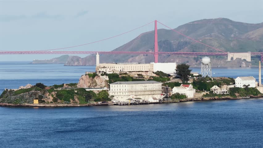 Alcatraz Island in San Francisco Bay, California, USA, with historic prison buildings, lighthouse and water tower, set against Golden Gate Bridge and coastal hills in calm scenic light. Aerial drone v