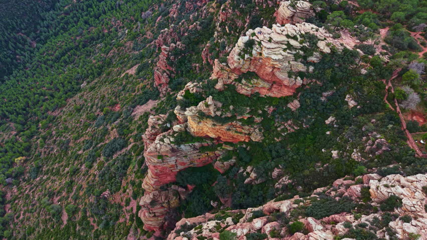 Aerial view shows rocky cliffs and greenery. The landscape features steep rock formations surrounded by trees. Sunlight shines on the terrain creating shadows.