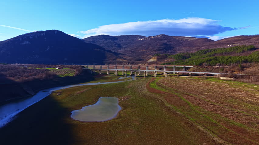 A bridge spans a dry riverbed surrounded by green hills and mountains under a clear blue sky. The landscape shows patches of water and land with low vegetation.