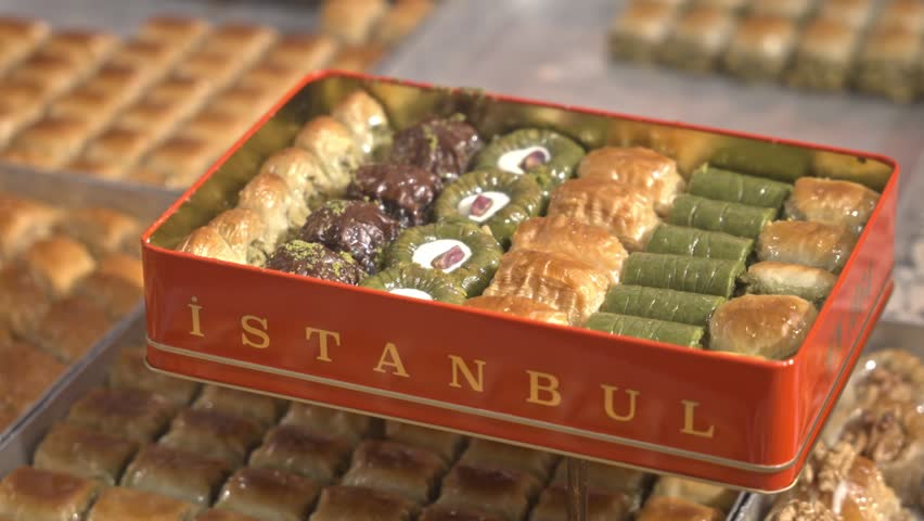 A mouth-watering 4K cinematic close-up of various traditional Turkish baklava types in a shop window. Features a red gift box labeled "ISTANBUL" among pistachio and walnut desserts.