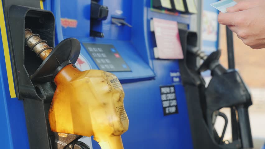 Woman paying for gasoline at outdoor gas station using credit card and touch screen display on pump, fueling vehicle with petrol pump nozzle, blue gas station, car fueling with credit card payment.
