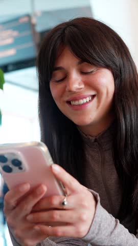 Beautiful young woman sitting in an airport terminal, smiling while using her smartphone and looking around, with a flight information display screen visible in the blurred background