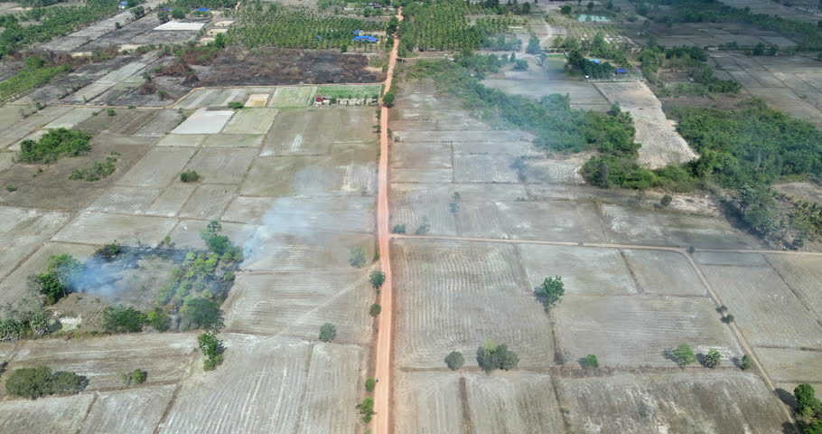 Backward aerial view of a straight red dirt road bisecting dry agricultural fields in rural with smoke from a field burn rising on the left.