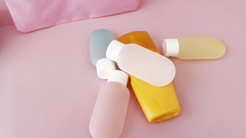 Woman preparing her skincare travel kit for a journey, placing shampoo, conditioner, lotion, and sunscreen bottles into a pink organizer on a matching background