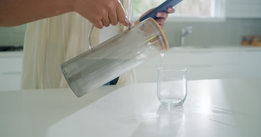 African American woman pouring water from jug into glass in kitchen while checking phone to hydrate. Pitcher, home, sunlit, bright, modern, minimal, lifestyle