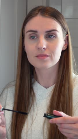 Young woman applying mascara while looking into mirror during daily beauty routine at home. Concept of makeup, cosmetics, selfcare, feminine lifestyle and natural beauty.