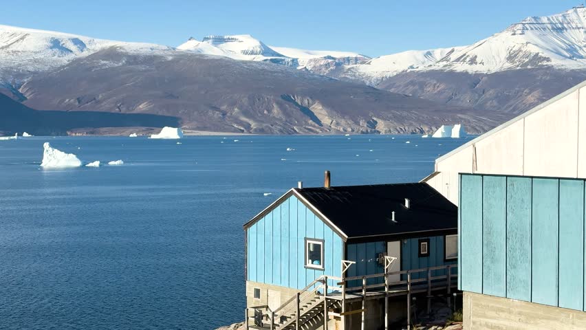Colorful houses sit on the rocks near the ocean in Greenland. Icebergs float in the water. The sun shines brightly. A few boats are seen near the shore. This location shows daily life by the sea.
