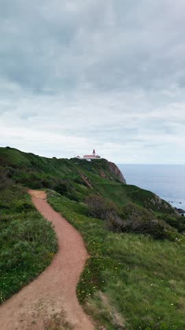 Establishing shot of a winding dirt trail along a grassy cliff overlooking the ocean, with Cabo da Roca lighthouse visible on a rocky outcrop near the water. Cascais, Portugal