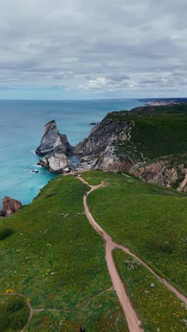 Drone aerial view of rugged coastal cliffs with a narrow dirt path leading toward the ocean, surrounded by green grass and rocky formations under a cloudy sky.