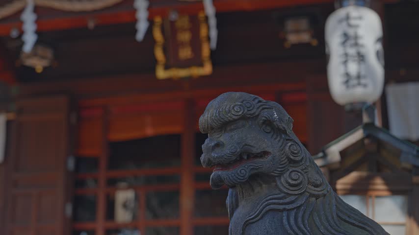 Close-up static shot of a dark stone komainu guardian statue, intricate curled mane detail, red-accented mouth, white paper lantern and warm wooden shrine hall softly blurred behind.