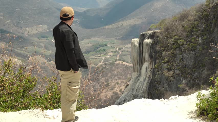 A young male traveler stands at Hierve el Agua in Oaxaca, Mexico, looking at the unique petrified waterfalls. The view shows mountains and valleys, capturing the beauty of this natural site.