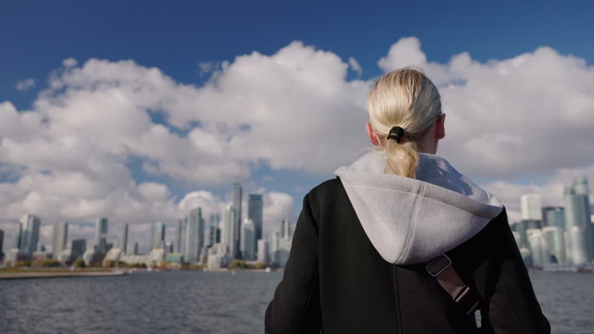 Young woman tourist looking at Toronto skyline from boat on Lake Ontario, back view with blonde hair enjoying urban cityscape and travel mood on waterfront.