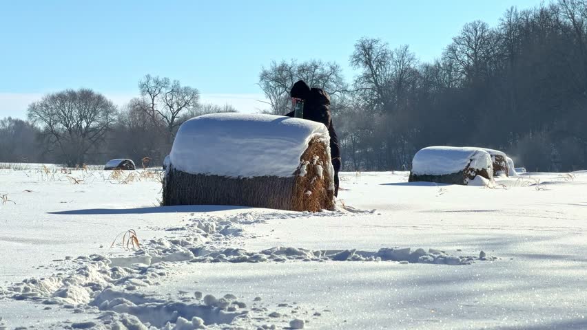 A person in dark clothing climbs onto a large, snow-dusted hay bale on a bright winter day.