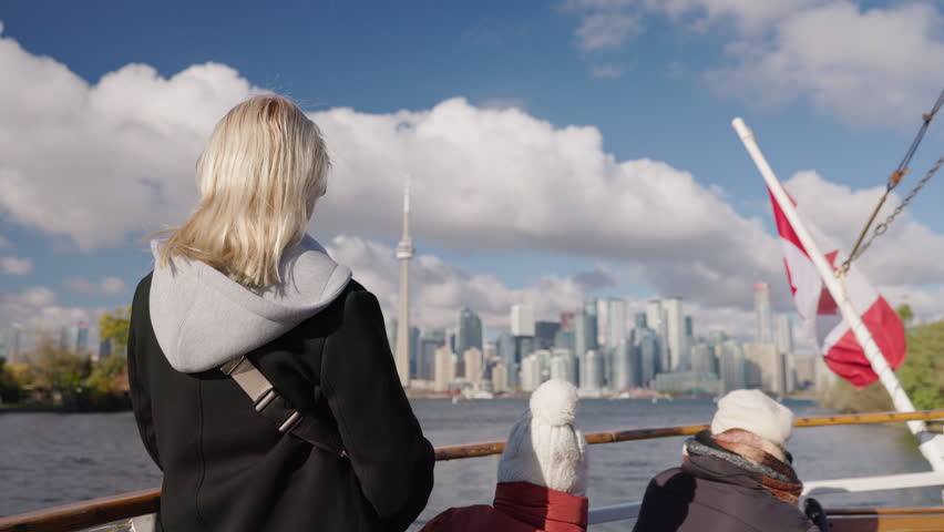 Young woman and two children passengers on boat looking at Toronto skyline from Lake Ontario, family travel scene with cityscape and Canadian flag in windy weather.
