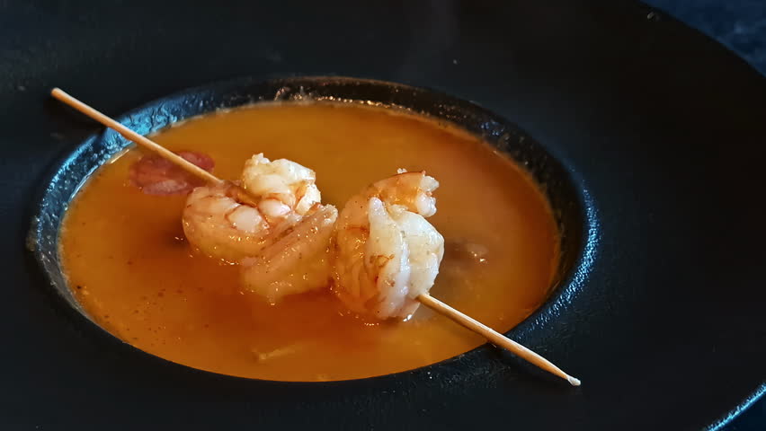 A rich, orange seafood soup served with a wooden skewer of two grilled shrimps.