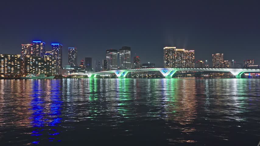 Wide static time-lapse of Harumi Bridge at night, green and blue decorative lighting reflecting on calm bay water, densely illuminated residential towers stretching across the skyline behind.