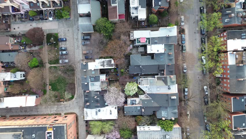 Residential spring grid blocks near S Paca St show mixed rooflines, narrow alleys, budding trees across dense residential rows. Varied shingles shape tight neighborhood textures Baltimore, MD. USA