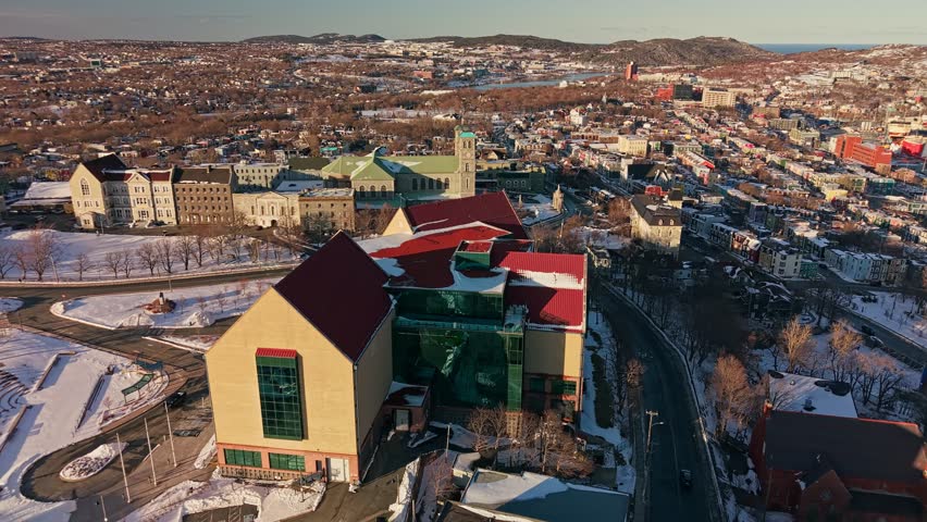 Aerial drone shot of The Rooms museum in St Johns Newfoundland with red rooftop snow and harbor ships visible beyond the colorful downtown