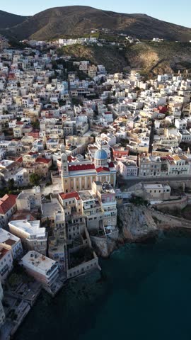 A spectacular vertical aerial drone video, perfectly formatted for social media, capturing a smooth orbit around the magnificent Agios Nikolaos church in Syros Ermoupoli, Greece. The stunning daytime footage highlights the elegant neoclassical mansions of the Vaporia neighborhood, the rugged coastal cliff, and the deep blue Aegean Sea.
