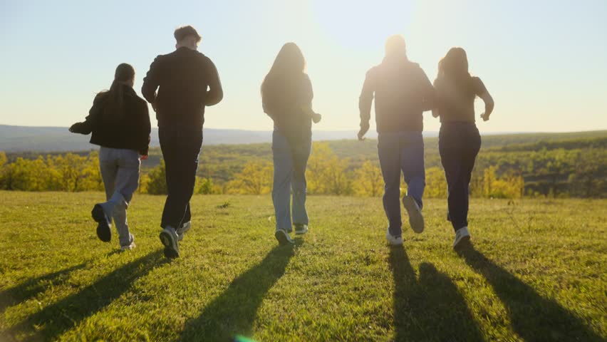 Rear view of a diverse group of five friends running through a grassy field during golden hour sunset, capturing a sense of freedom and unity.