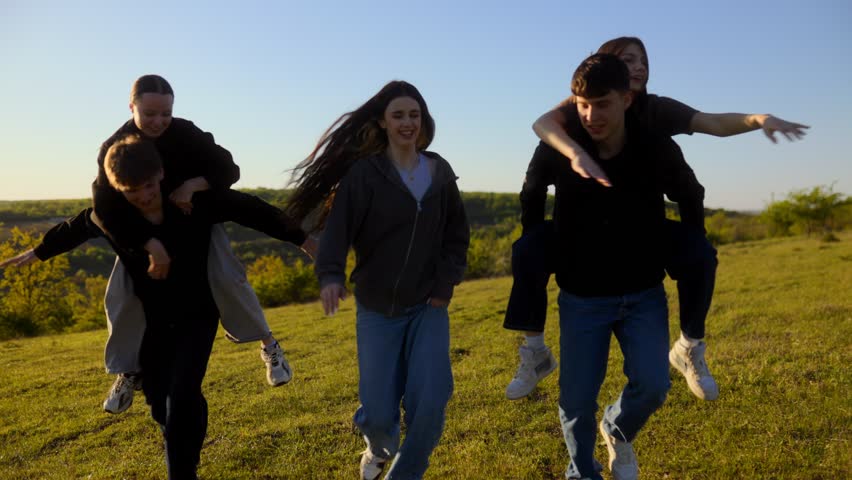 Group of five happy friends walking toward the camera with piggyback rides, laughing and playing in a green meadow during a beautiful warm sunset.
