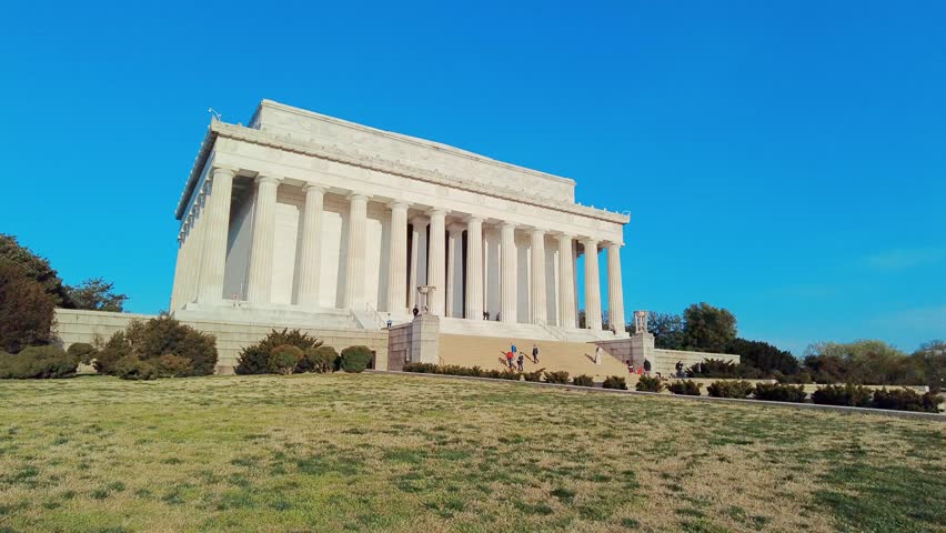 Washington DC. A clear day with a clear blue sky and a grassy hill in the foreground. The Lincoln Memorial is a neoclassical structure with a series of white columns and a central pediment.