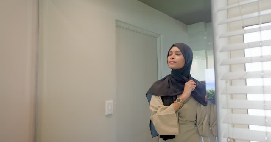 African American woman in beige wrap-top pulling hijab up, getting ready at home mirror, copy space. Headscarf, reflection, grooming, styling, drape, bathroom, editorial