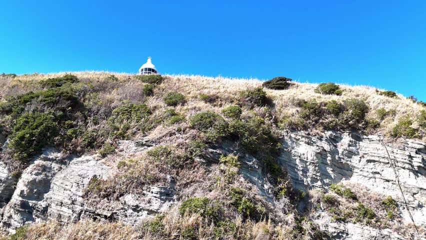Low angle aerial shot looking up at Tsurugisaki Lighthouse standing tall on a cliff edge against a blue sky.
